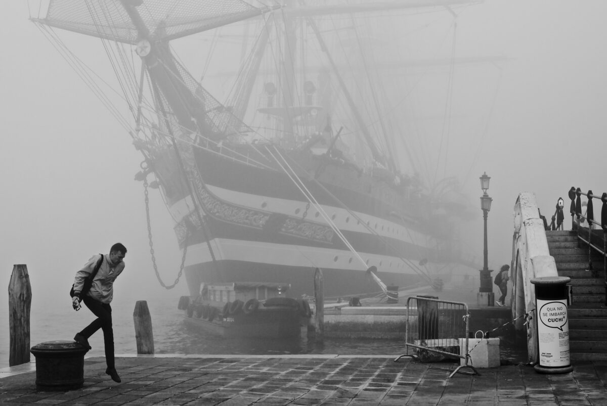 Tall ship Amerigo Vespucci in the morning fog in Venice, with man stepping down from mooring bitt in the foreground.